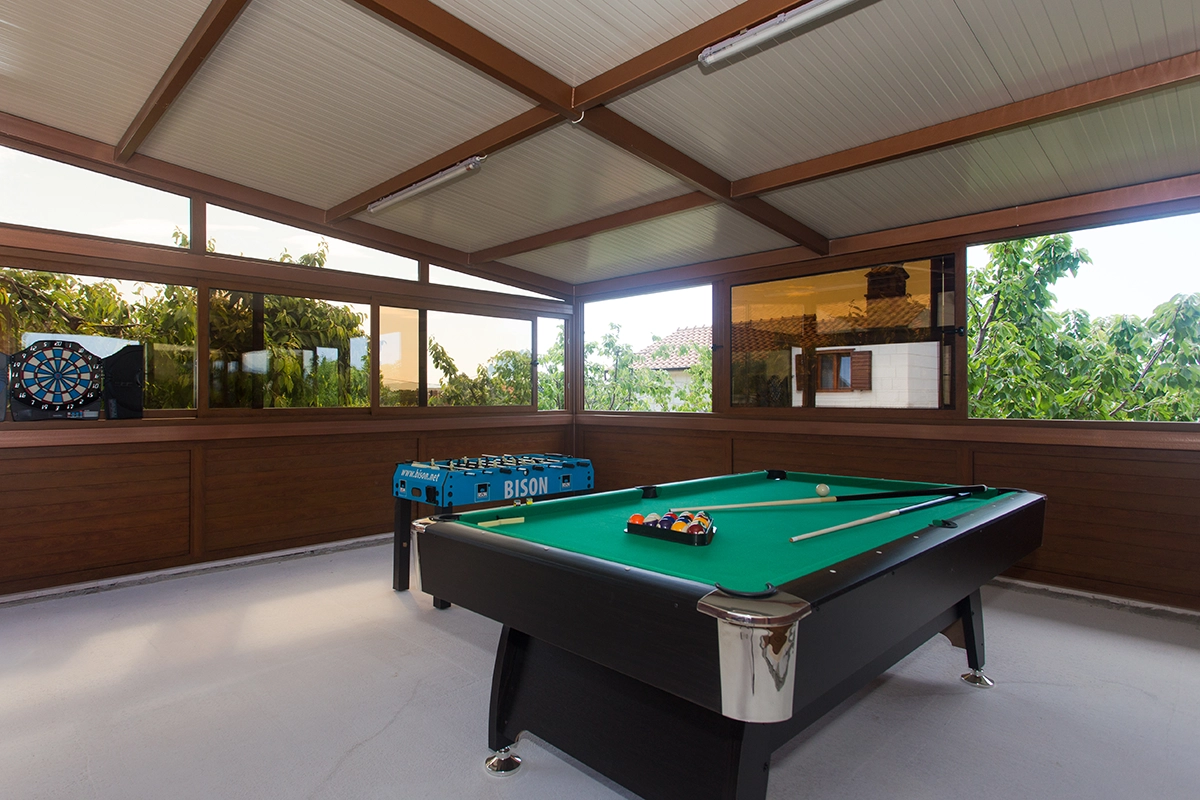 Enclosed outdoor wooden space designed for entertainment, equipped with a dartboard, foosball table, and billiards, picture from different angle.